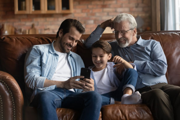 A grandparent, his son, and his grandchild sit on a couch and watch a funny video on the son's mobile.