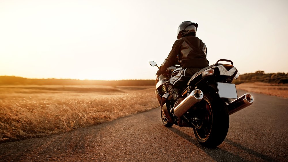 A motorcyclist parks at the side of the road and watches the sun set over a large wheat field