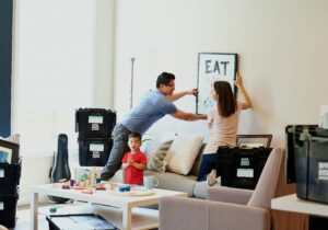 A couple mounting a picture frame on the wall of their living room. Boxes containing possessions are stacked around the room.