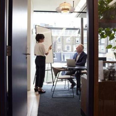 A businesswoman uses a whiteboard to present ideas to a businessman in a small meeting room.